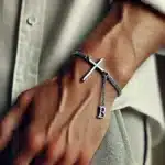 DALL·E 2024-11-25 20.26.42 – A close-up of a man’s wrist wearing a bracelet featuring a horizontal cross with a delicate silver chain. There is a small dangling letter ‘B’ attache